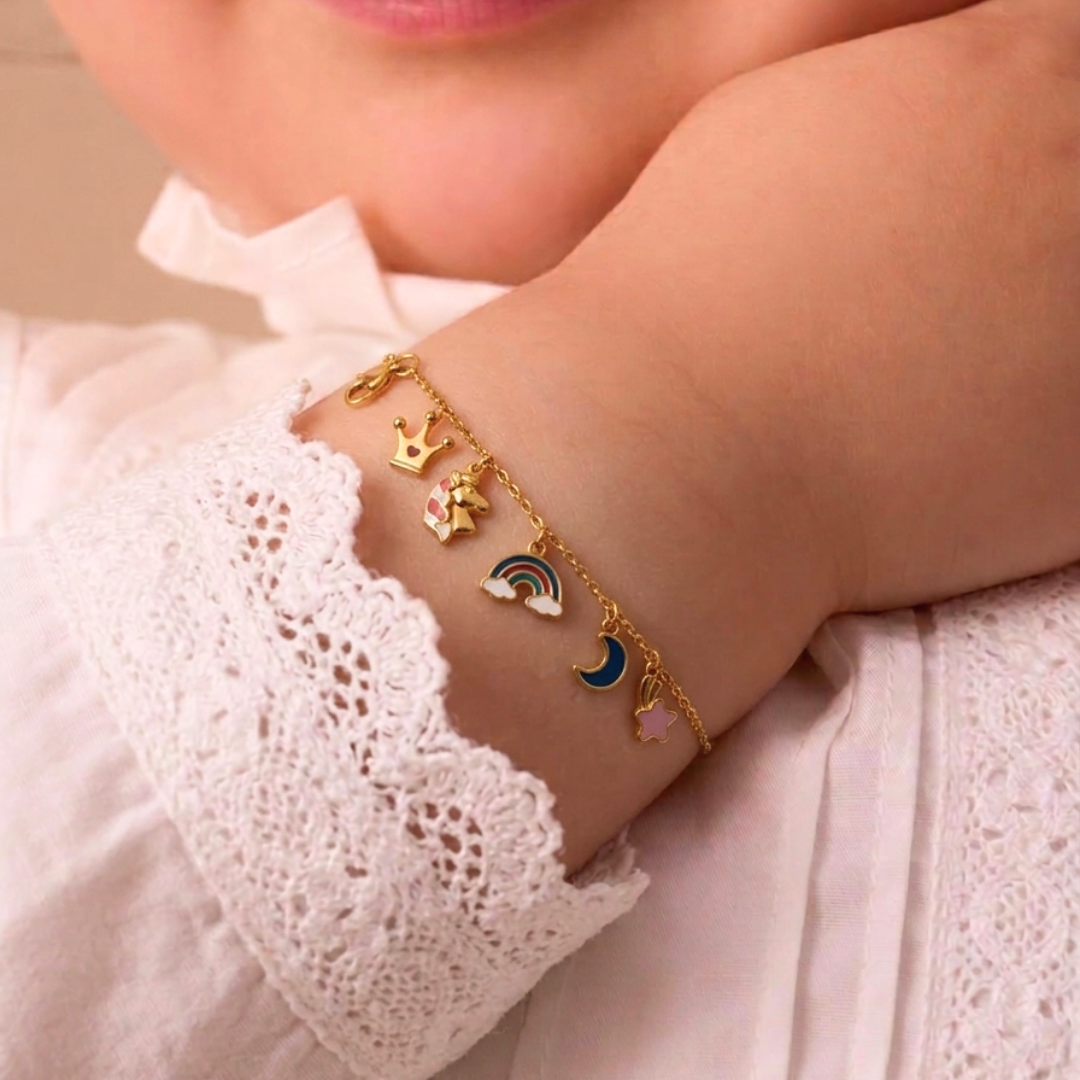 Gold charm bracelet with crown, star, moon and rainbow enamel charms, worn on a baby's wrist.
